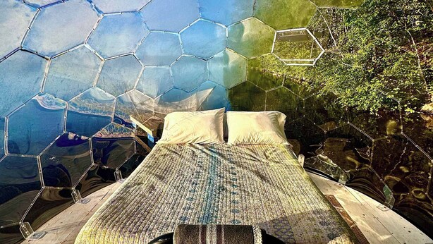 Ein Zimmer mit einer sechseckigen Glasdecke, einem Bett mit karioter Decke und Blick ins Grüne.