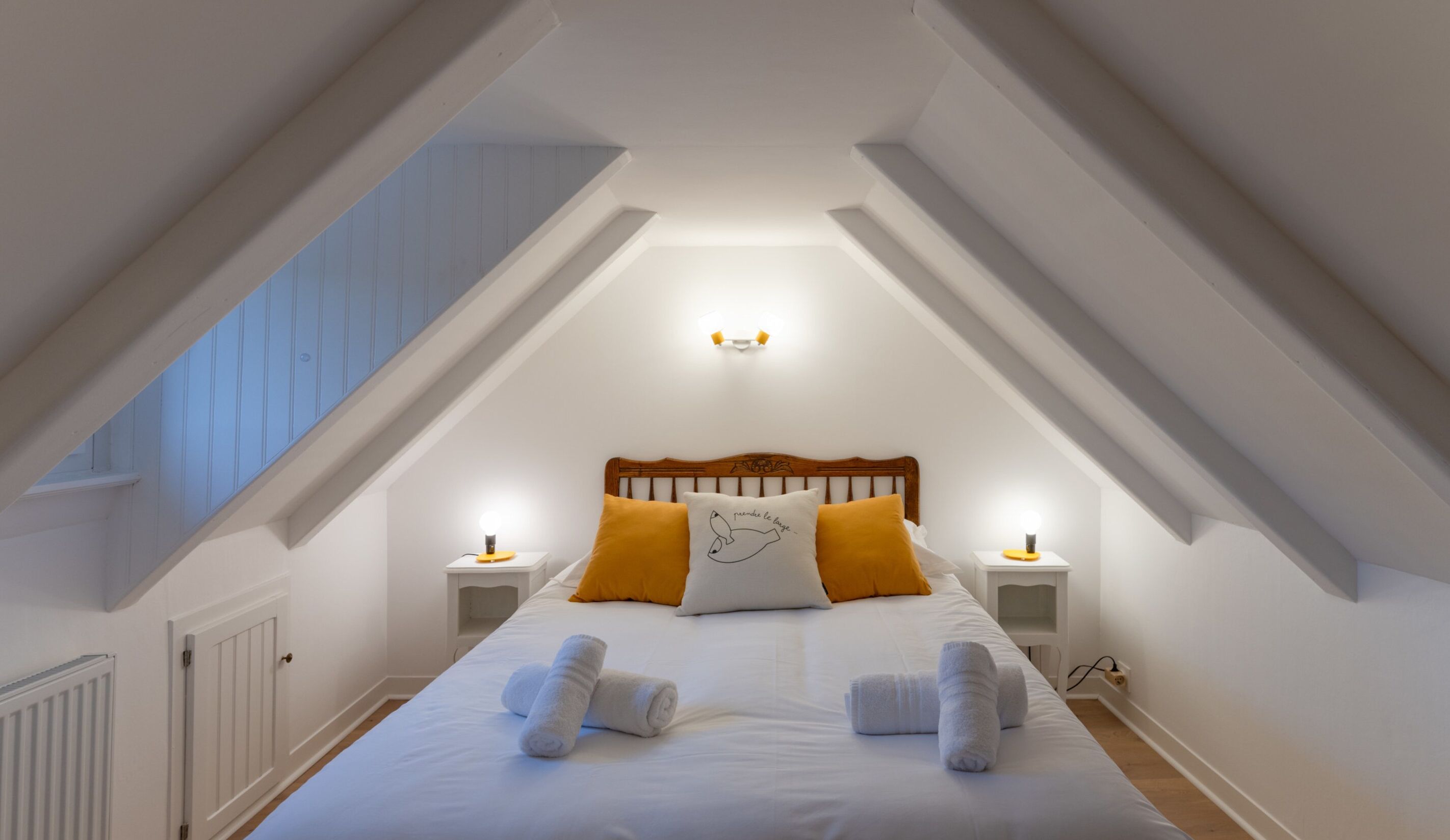 A bedroom with a sloped ceiling, a wooden headboard, two bedside tables with lamps, a bed with white bedding and two pillows, and a window with blue shutters.