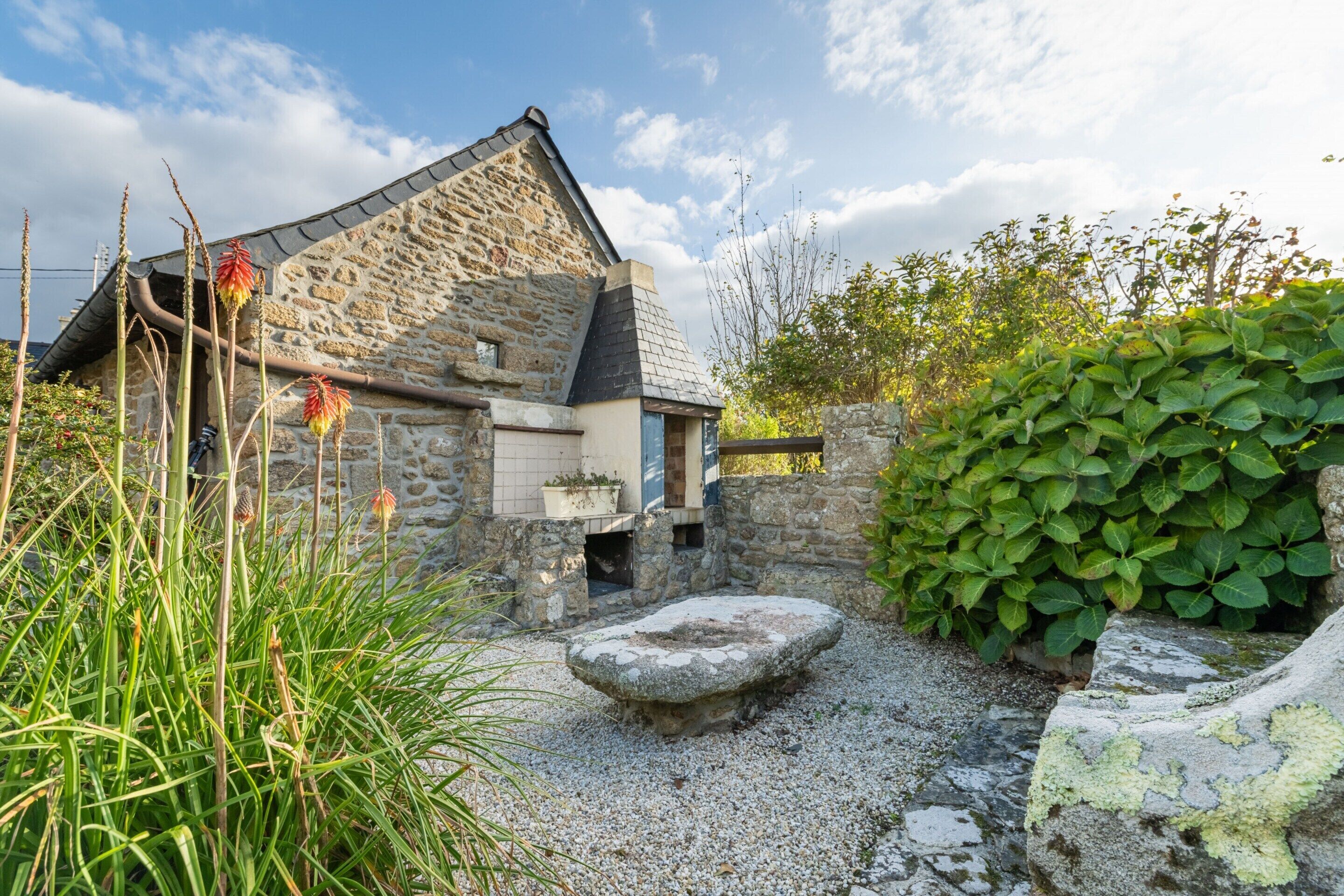 A stone building with a sloped roof, surrounded by gravel, plants, and a stone bench.