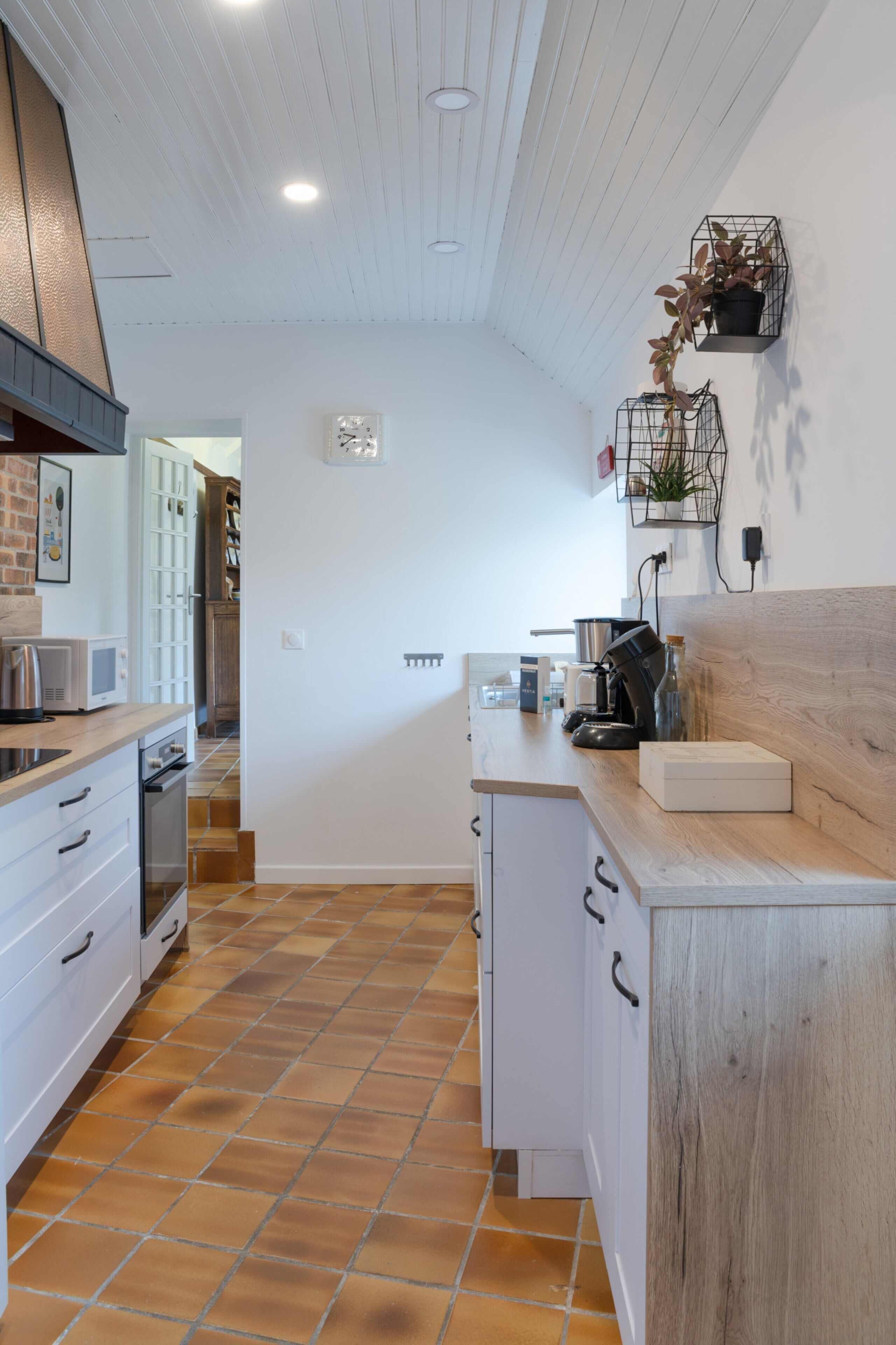A modern kitchen with white cabinets, a wooden countertop, and a tiled floor. There are two wall-mounted shelves with plants and a coffee machine on the counter.