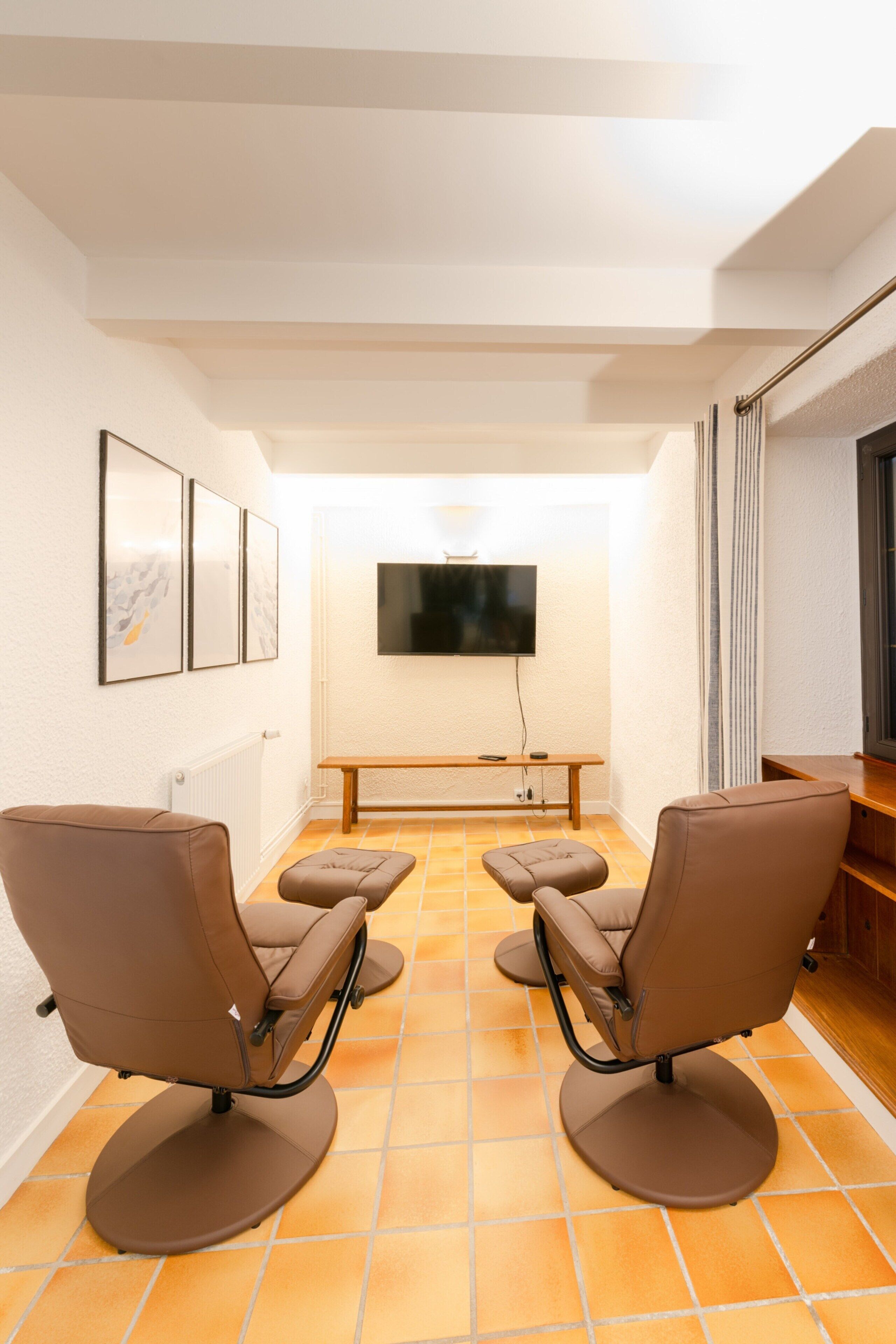 Ein Zimmer mit zwei braunen Stühlen, einer Bank, einem flachbildfernseher an der Wand und einem gefliesten Boden.