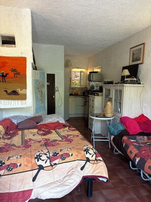 A compact room with a bed, a small table, a chair, a kitchenette, and a painting on the wall. -  1-bedroom apartment in charming Liscia beach :4 pers max: 2 weeks +3rd free (Calcatoggio)
