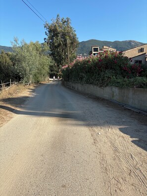 A rural road with a dirt path, surrounded by vegetation and a building with a sloped roof. -  1-bedroom apartment in charming Liscia beach :4 pers max: 2 weeks +3rd free (Calcatoggio)
