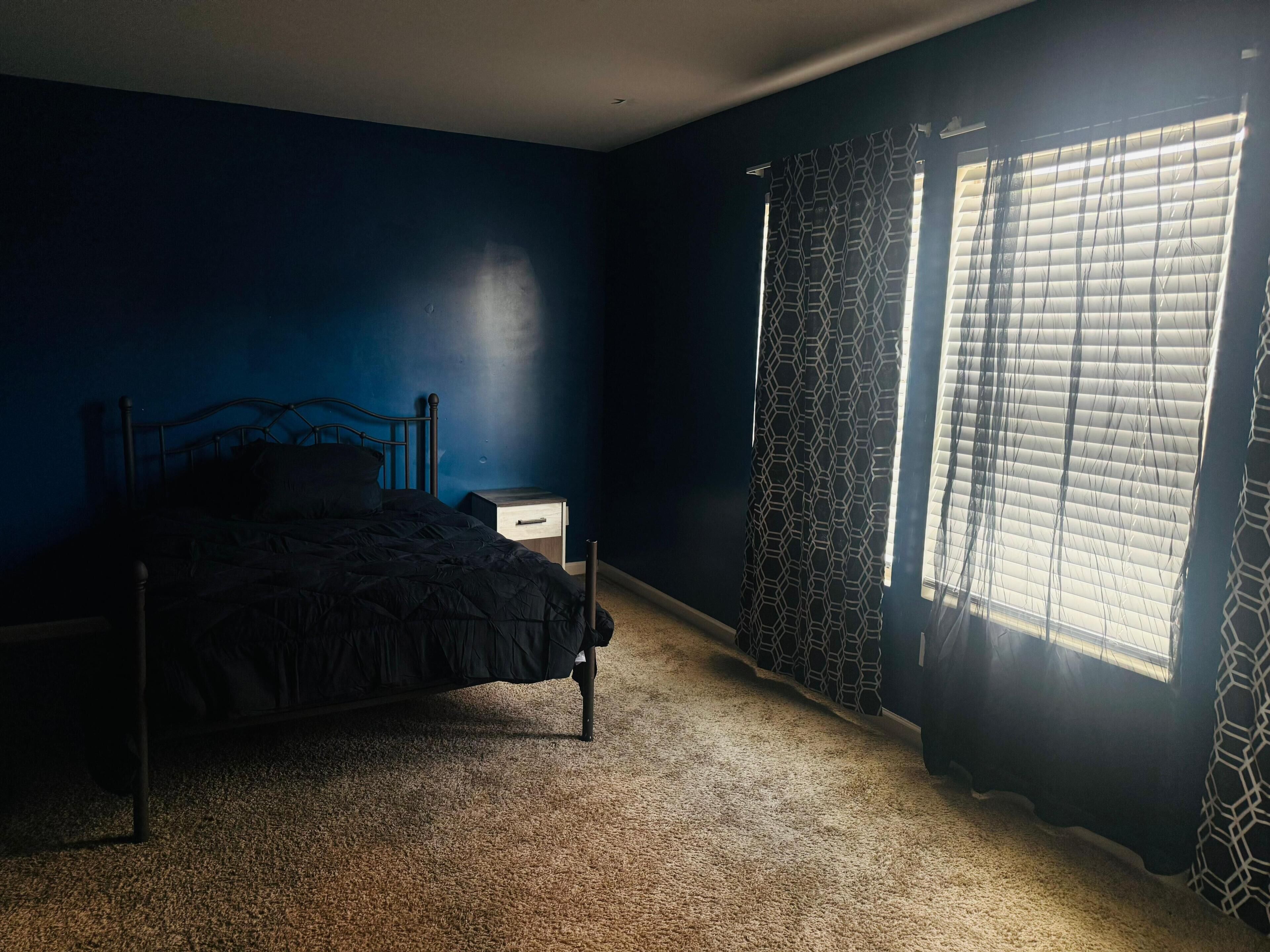 A bedroom with a bed, a nightstand, a window with patterned curtains, and a dark blue wall.