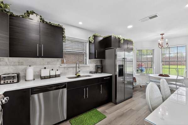 A modern kitchen with dark cabinets, stainless appliances, and a dining area with a white table and chairs. - NEW! Central Frisco Family Getaway (Frisco)