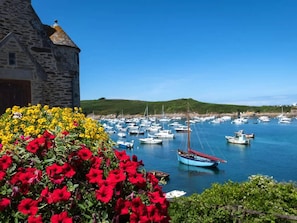 Un paysage côtier avec un bâtiment en pierre, des fleurs aux couleurs vives et un port de plaisance où sont amarrés différents bateaux.