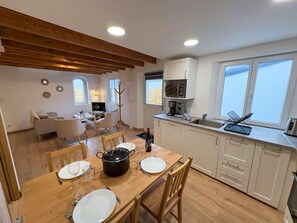 Une cuisine moderne avec des meubles blancs, une table à manger pour quatre personnes et une grande fenêtre qui laisse entrer la lumière naturelle.
