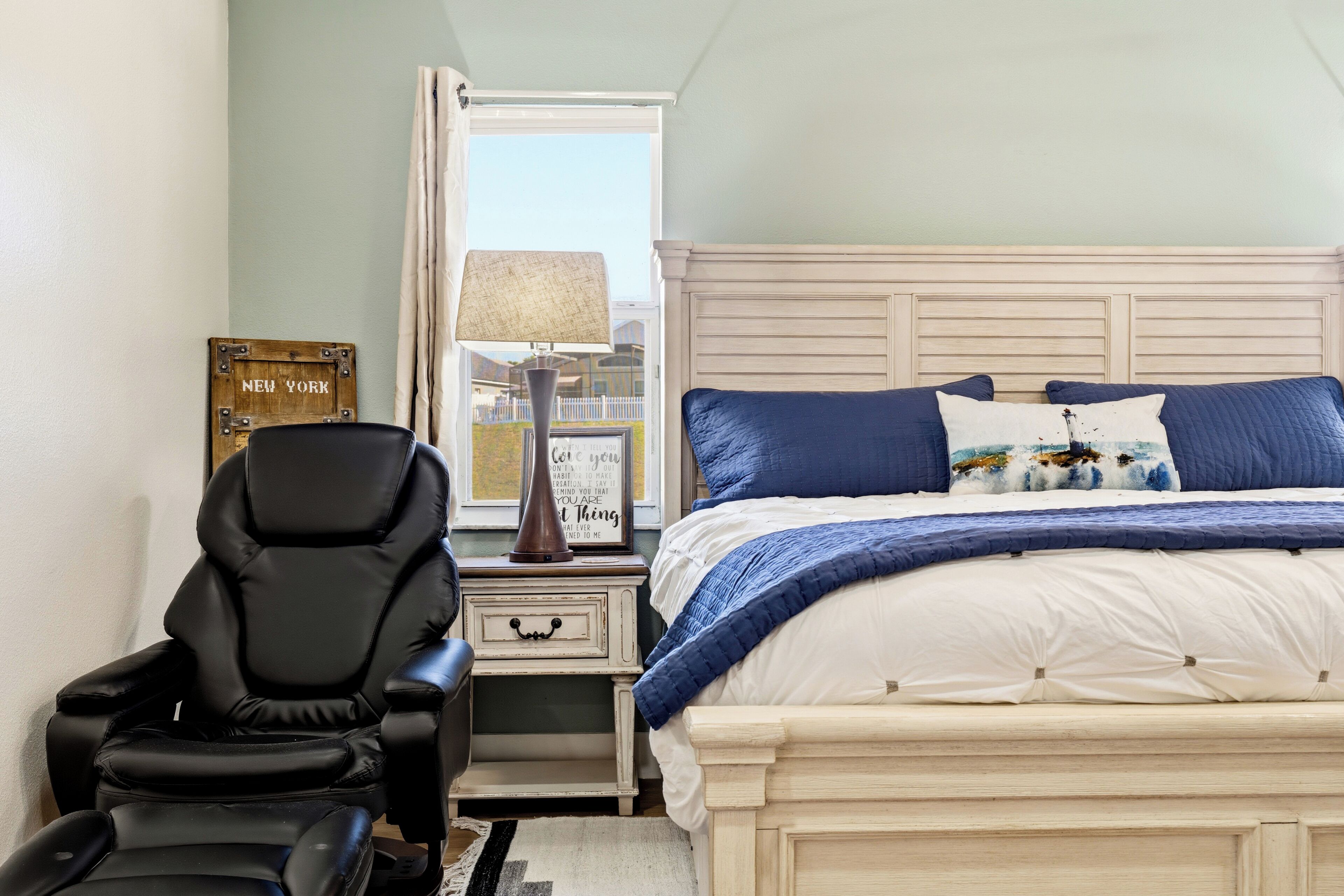 A bedroom with a bed, a chair, a nightstand, a lamp, a window, and a wall with a sign.