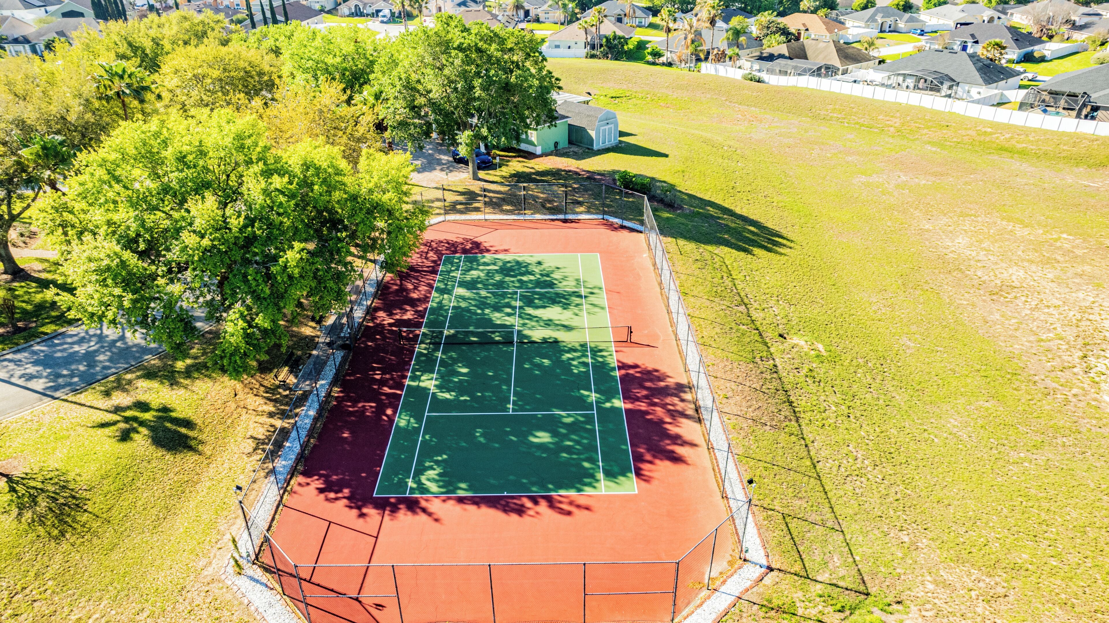 An aerial view of a tennis court surrounded by trees and residential buildings.
