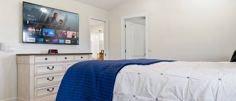 A bedroom with a bed, a dresser, a flat-screen TV mounted on the wall, and a door leading to another room.