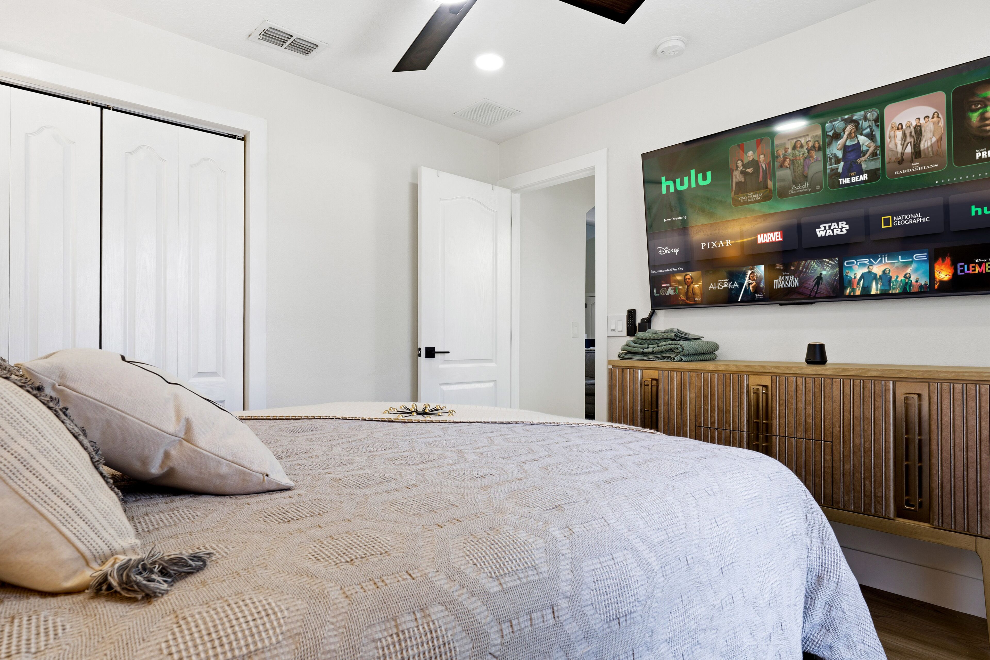 Un dormitorio con una cama, un televisor instalado en la pared mostrando un servicio de streaming, un armario de madera y una puerta que da a otra habitación.