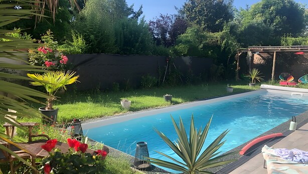 Ein Swimmingpool mit einem umlaufenden Garten, einer Pergola und Blick auf Bäume.