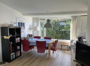 A dining area with a table, chairs, a bookshelf, a television, and a window with a view of greenery. - 6-person Bungalow in Burgerbrug (Nieuwvliet)