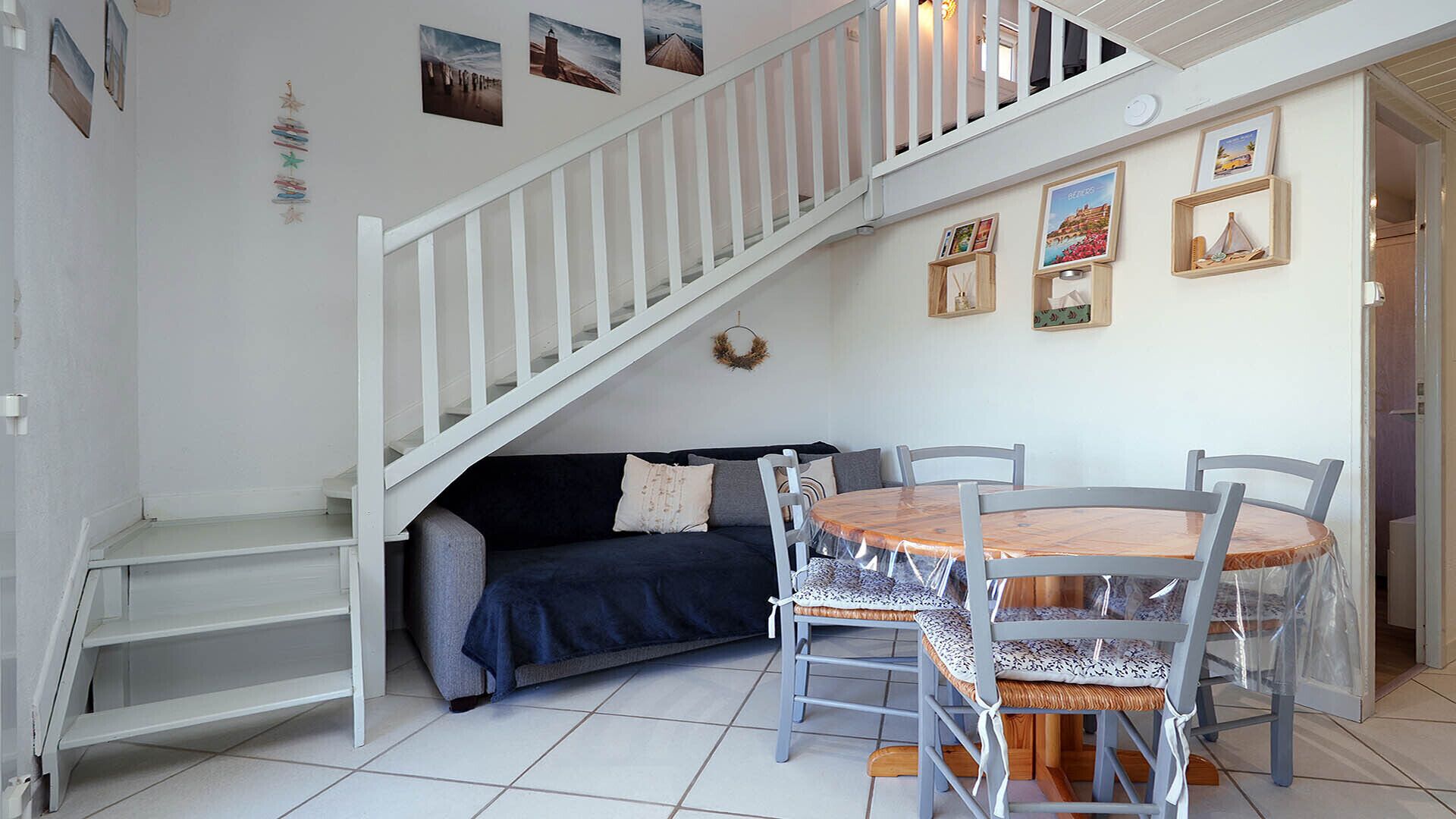 A staircase leading to an upper level, a dining area with a round table and chairs, a sofa with cushions, and a wall decorated with framed pictures.