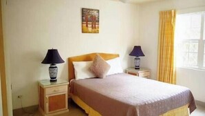 A bedroom with a bed, two bedside tables, a nightstand, a window with curtains, and a painting on the wall. - Shades Apt 1 – Spacious Two-Bedroom Garden Apartment Near the Pool (Saint James)