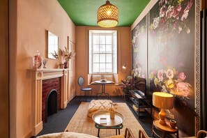 A cozy room with a fireplace, a round table, a lamp, a piano, and a floral mural.