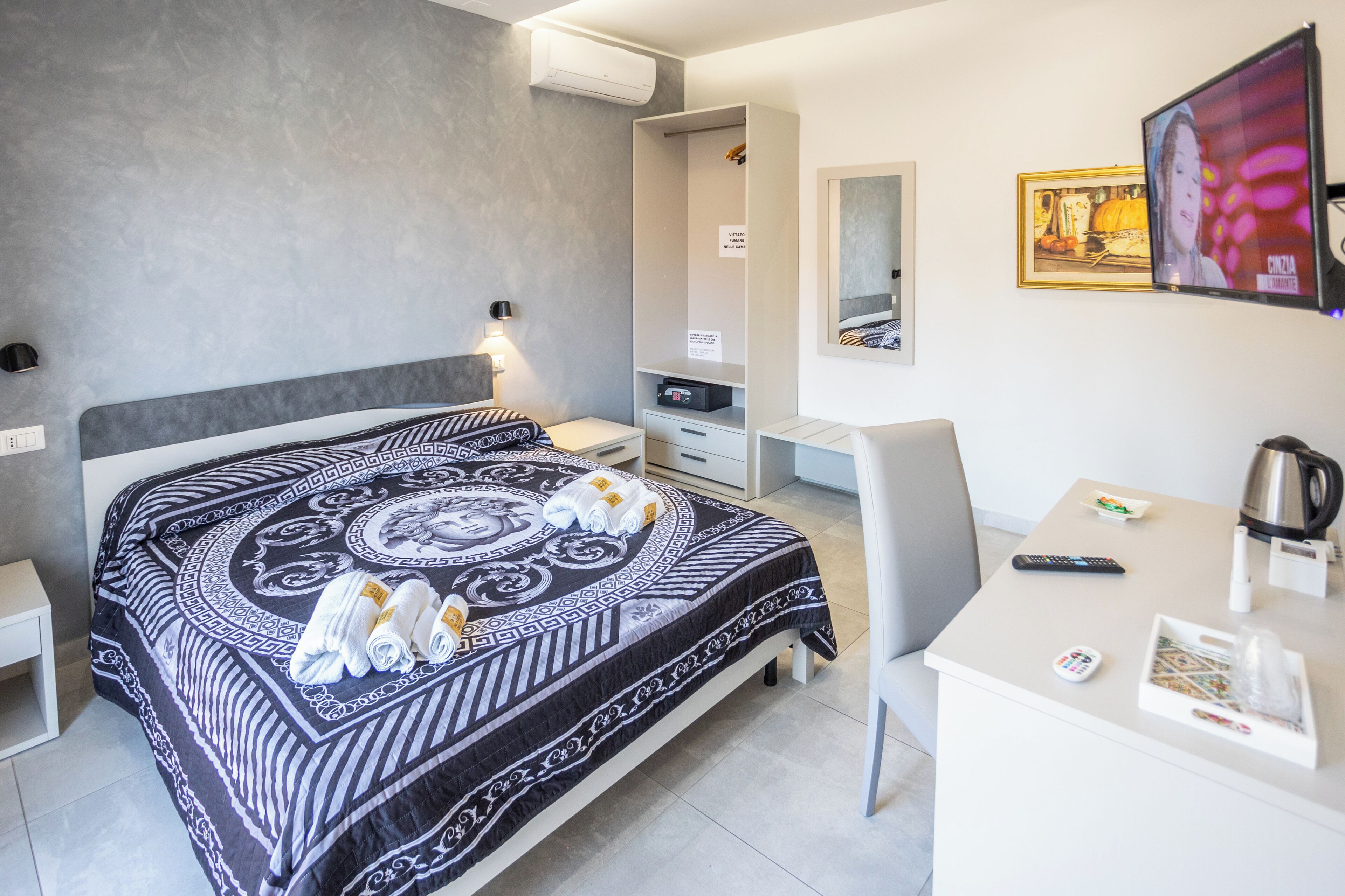 Habitación de hotel moderna con una cama grande, un escritorio con hervidor, una silla y un televisor montado en la pared.
