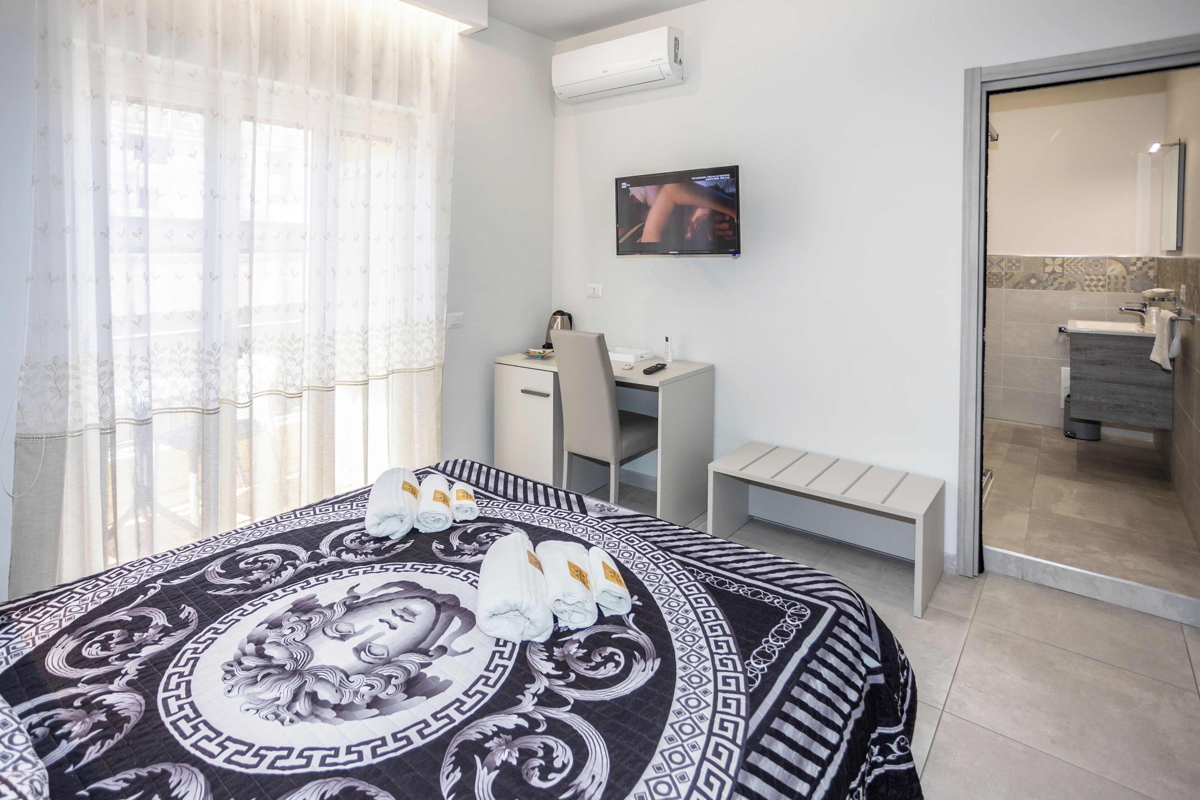 A hotel room with a bed, a desk, a chair, a TV, and a bathroom visible through a glass door.