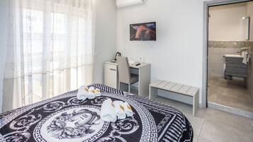 A hotel room with a bed, a desk, a chair, a TV, and a bathroom visible through a glass door.