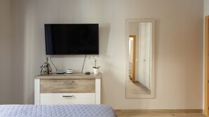 Quarto com cama, televisão na parede, cômoda, espelho e porta que dá para outro espaço.