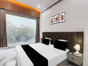 A hotel room with a large window, a bed with black and white bedding, a nightstand with a lamp, and three framed pictures on the wall. - Super Collection O Quila Road (Lucknow)