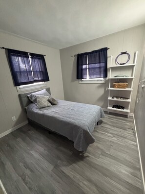A bedroom with a bed, two windows with curtains, a white shelf with various items, and a grey floor.