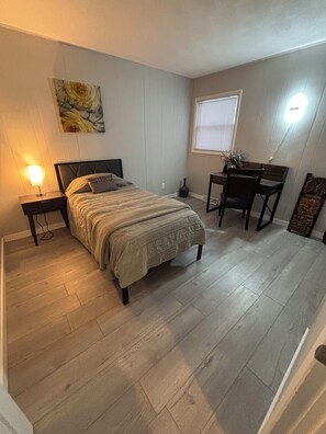 A bedroom with a bed, bedside table, lamp, desk, chair, window, and artwork on the wall.