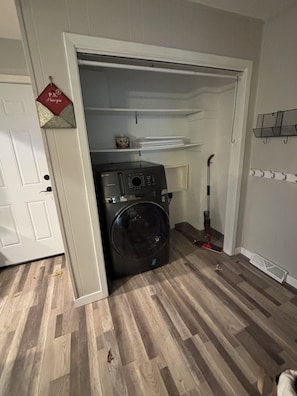 A laundry room with a washing machine, a mop, and a shelf with folded items.