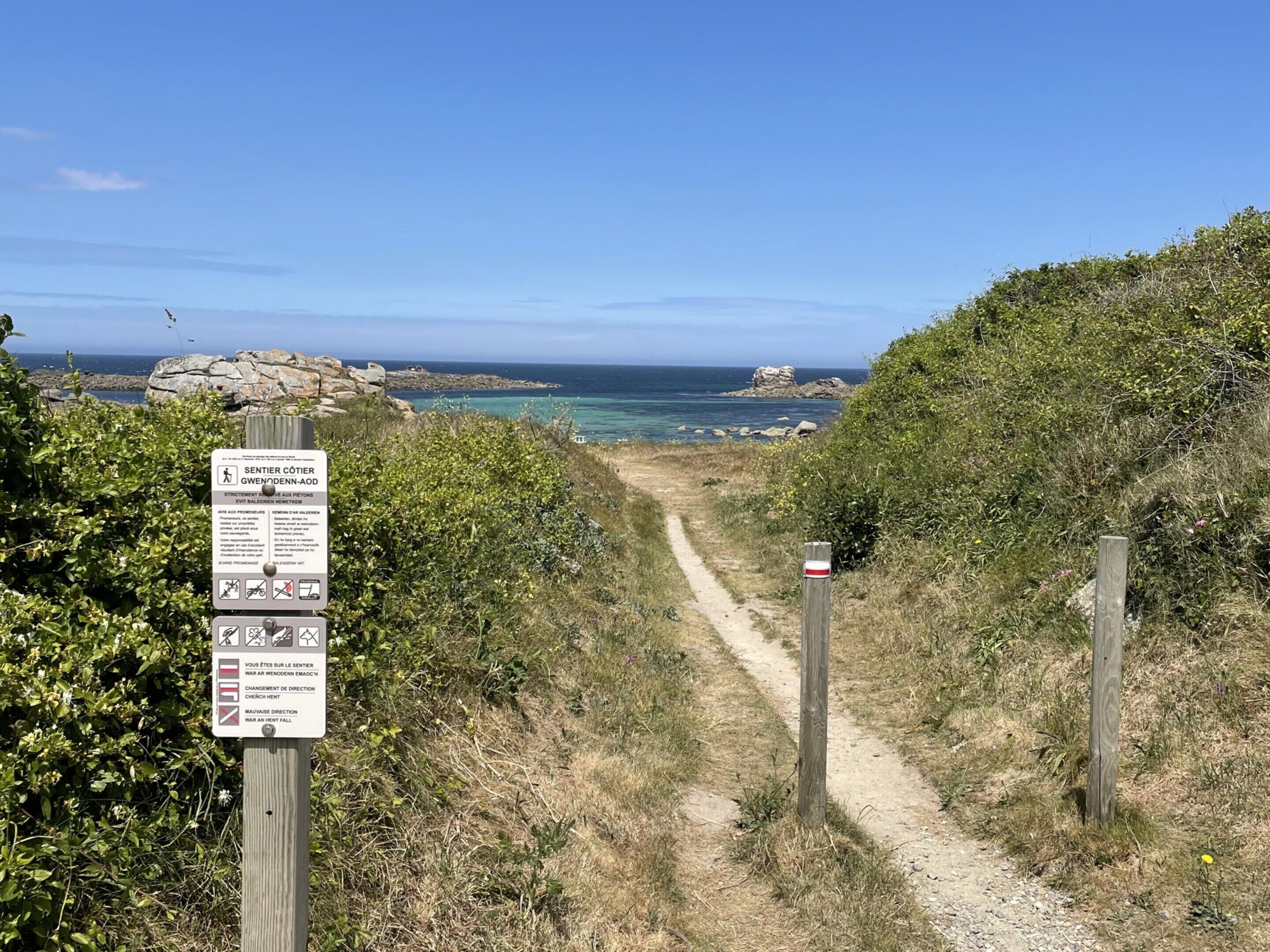 Un sentier de randonnée avec un panneau indicateur, une côte rocheuse et un ciel bleu dégagé.