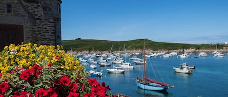 Escena costera con un edificio de piedra, un puerto lleno de barcos y flores coloridas en primer plano.