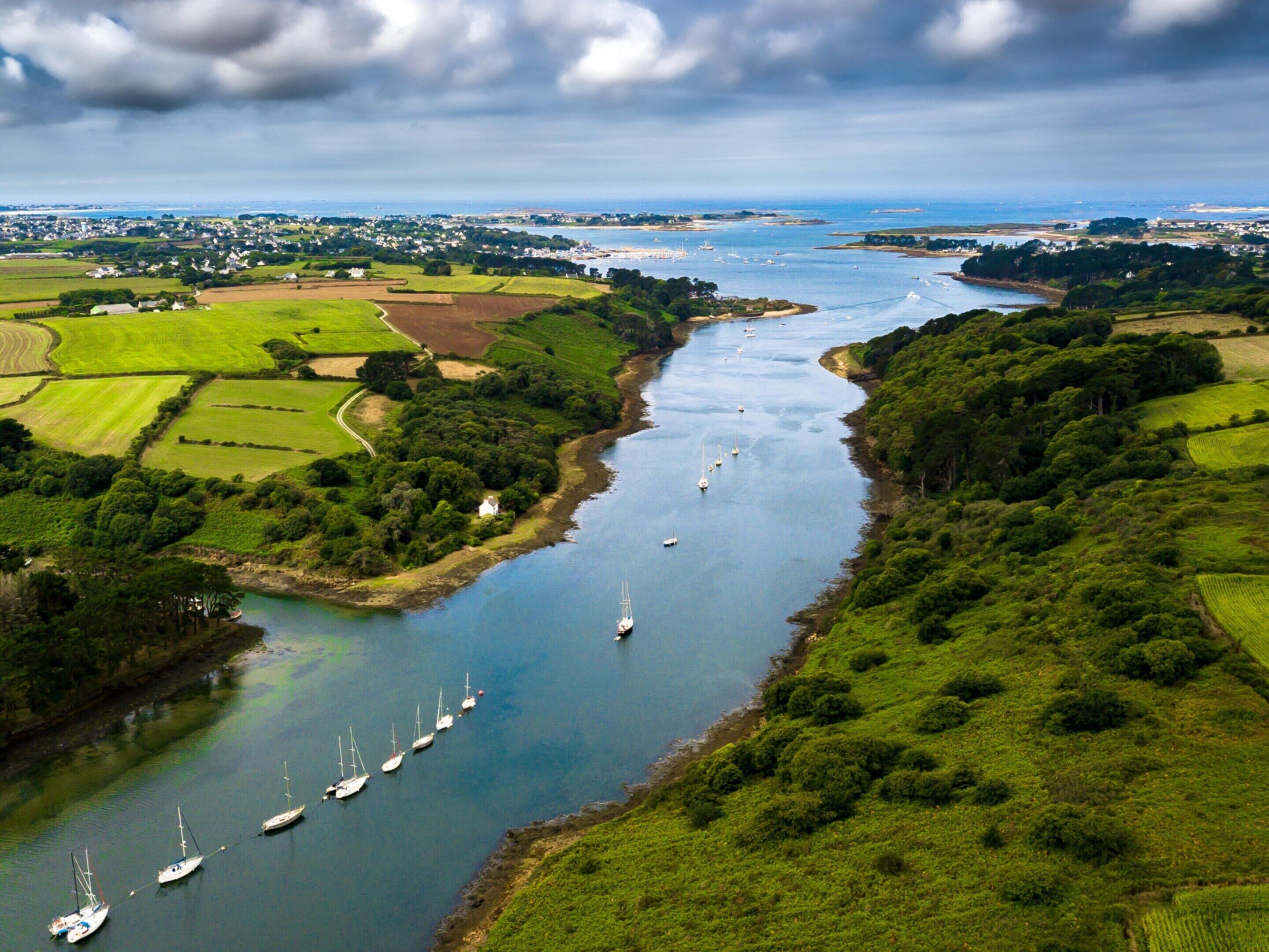 Une rivière où naviguent des bateaux, bordée de champs verdoyants et avec une ville qui se profile à l’horizon.