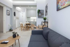 Ein modernes Wohnzimmer mit einem grauen Sofa, einem Holzesstisch und Stühlen, einer Küche im Hintergrund und einer kleinen Pflanze an der Wand.