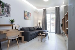 Ein modernes Wohnzimmer mit einer Couch, einem Schreibtisch, einem Sessel, einer Lampe und Blick durch ein großes Fenster.