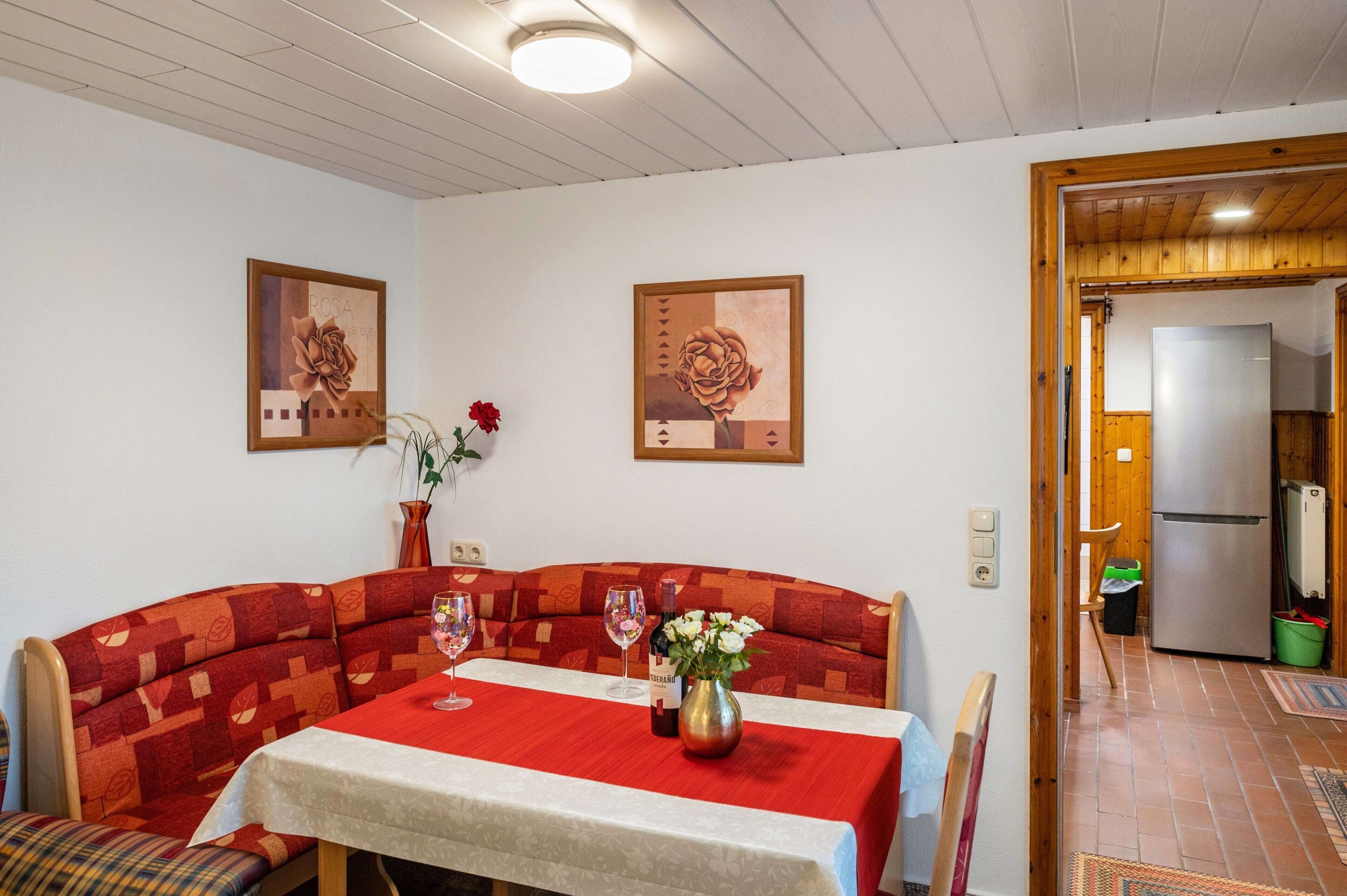 A dining area with a red and white table, two framed floral paintings, and a wooden door leading to a kitchen.