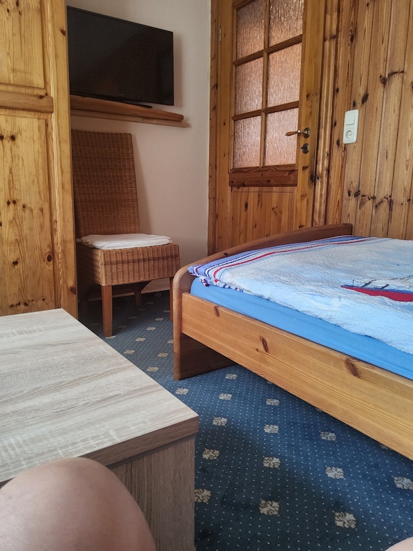 Una habitación de madera con una cama, una silla de mimbre, un televisor en la pared y un armario de madera.