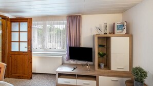 Ein Zimmer mit Holztür, einem fenster mit Vorhängen, einem Fernseher auf einem Holzschrank, einem Schrank mit Glastür und einer kleinen Pflanze.