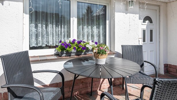 Eine Terrasse mit einem runden Tisch, vier Stühlen, einem fensterartigen Öffnung mit Vorhängen und einem Blumentopf mit Blumen.