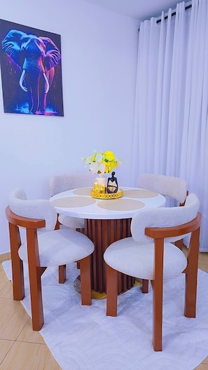 A dining area with a round table, four chairs, a vase with flowers, and a wall-mounted elephant painting. - Modern Coastal Apartment - Lift/Elevator Access,7 mins from beach, 24hr security (Mombasa)