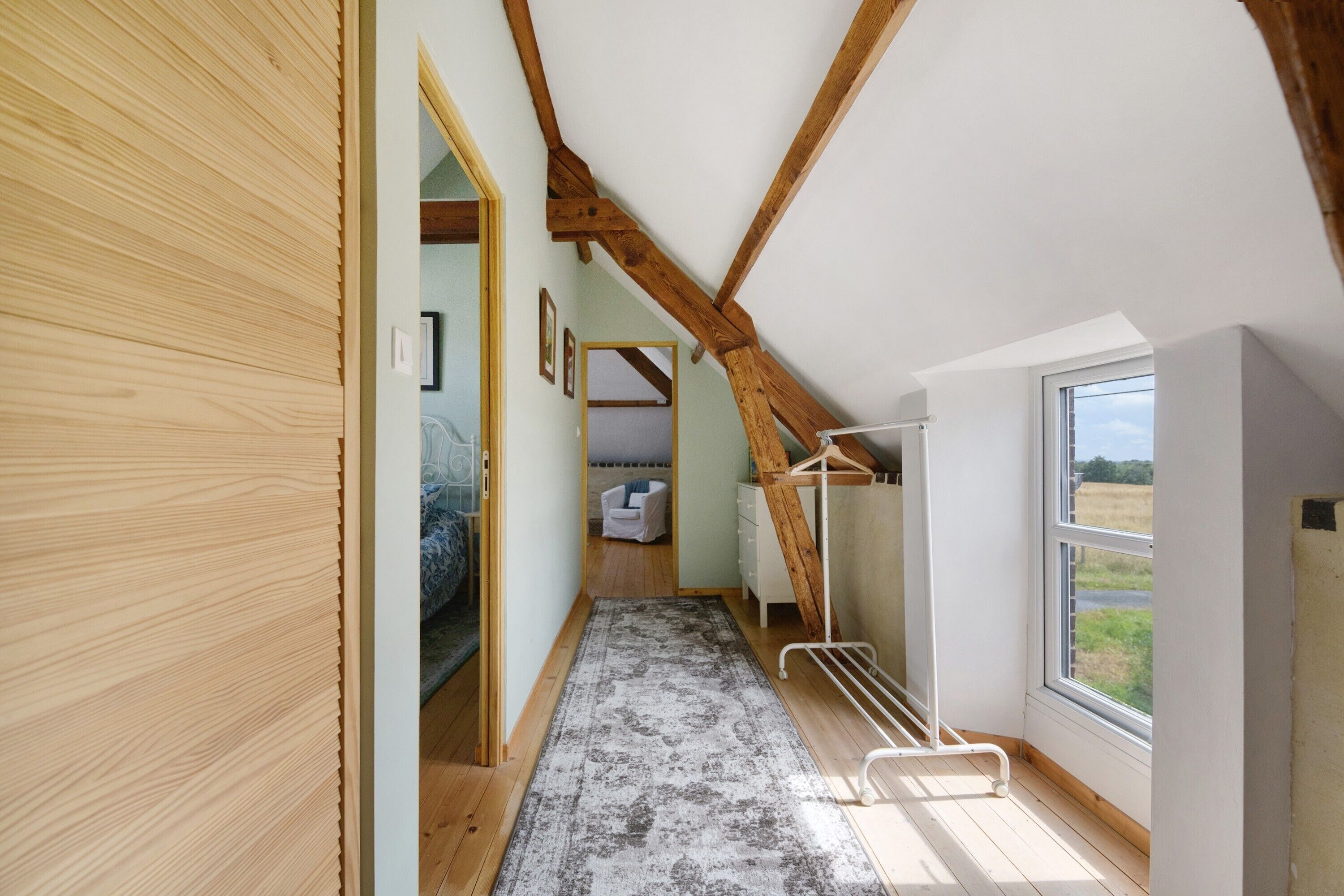 Un pasillo estrecho con vigas de madera, una cama, una silla y una ventana con vistas al exterior.