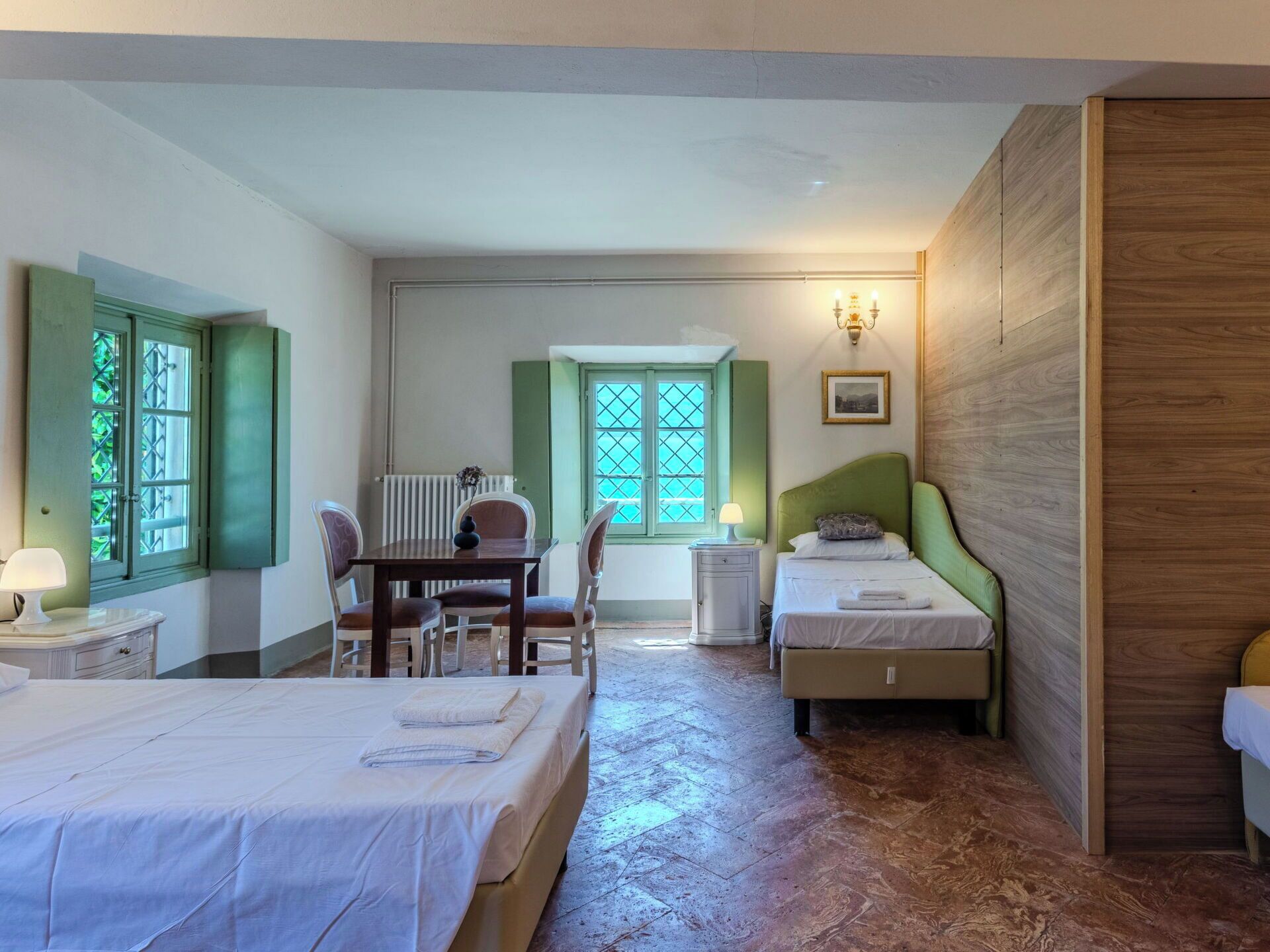 A bedroom with two beds, a dining table, chairs, a radiator, and a window with shutters.
