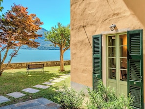 Eine Bank am See, ein Baum und ein Gebäude mit Fenster und Fensterläden.