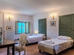 Ein Hotelzimmer mit zwei Betten, einem Sessel, einem Tisch, einem Heizkörper, einem Fenster mit Rollläden und Wandleuchten.