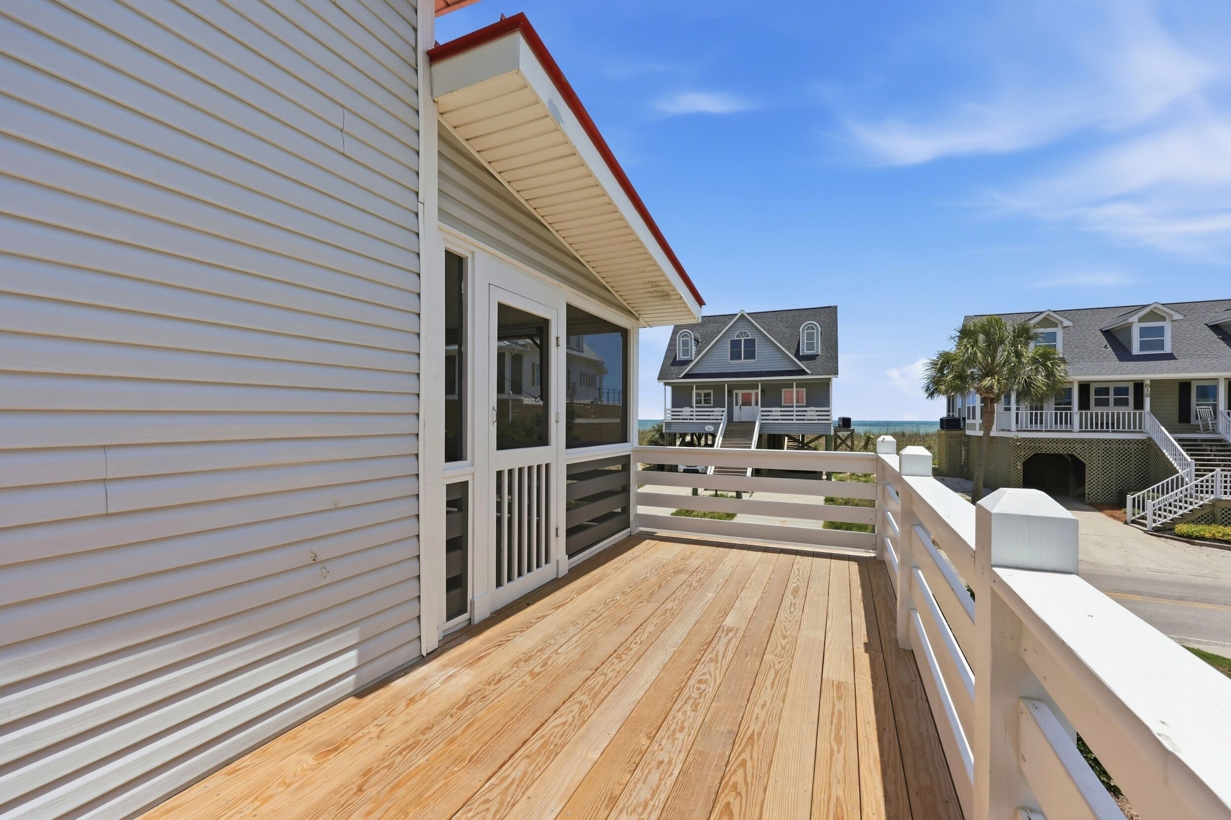 Une terrasse en bois avec une rambarde blanche, un espace couvert doté de portes vitrées et une vue sur un immeuble résidentiel avec balcon.