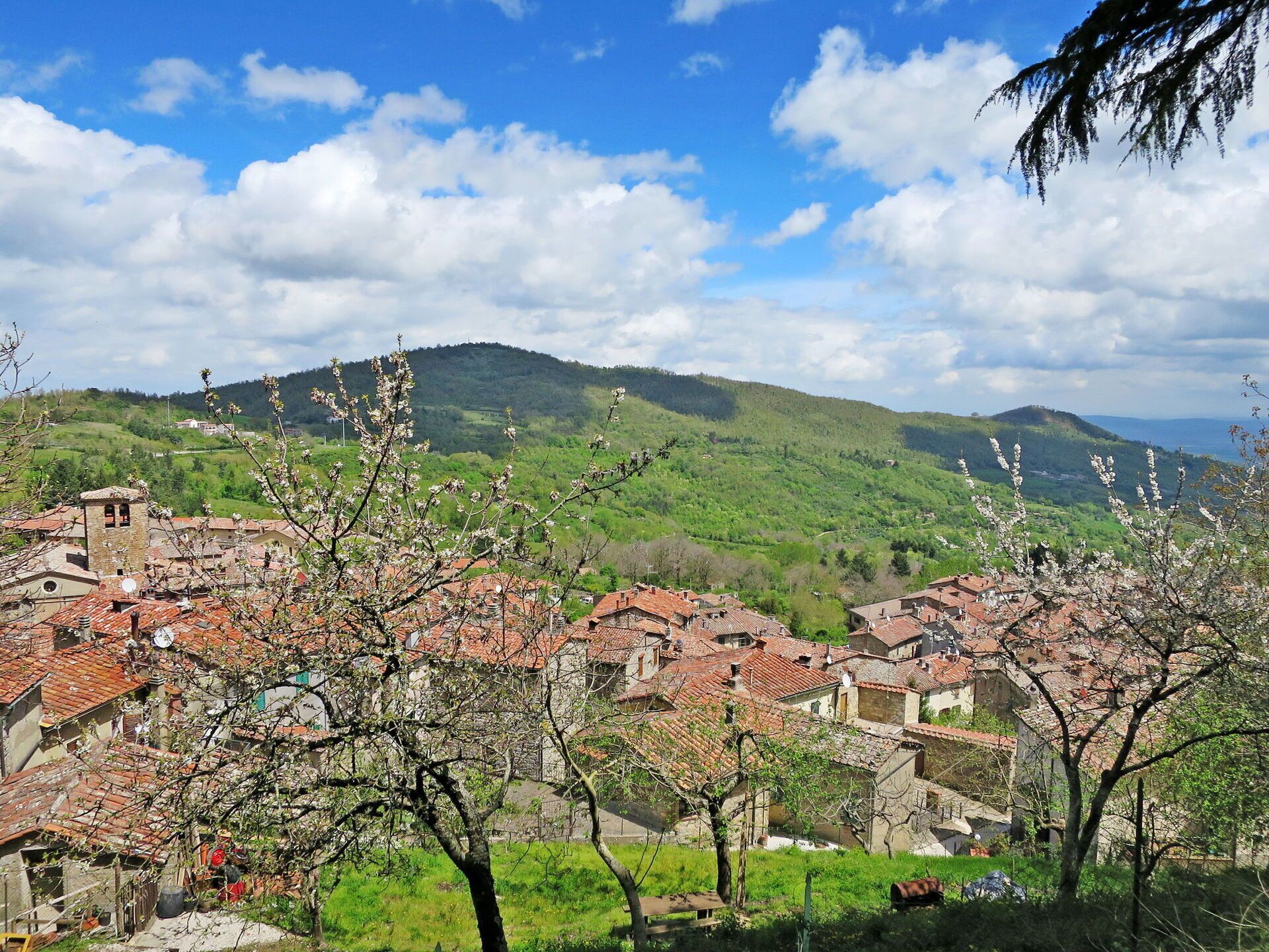 A village with traditional red-tiled roofs, surrounded by lush green hills and a clear blue sky.