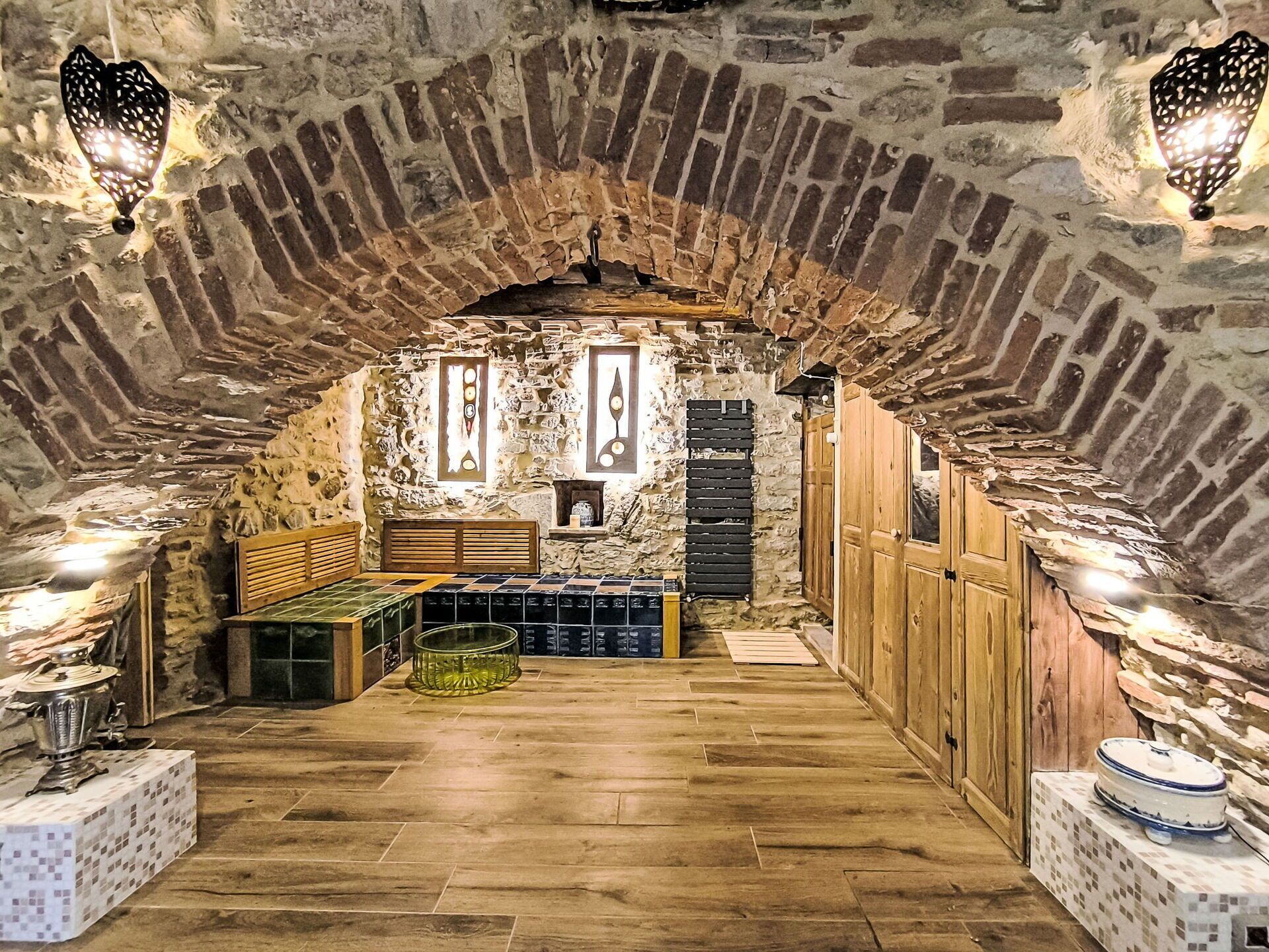 A stone-walled room with a wooden bench, a tiled floor, and a shelf with bottles.