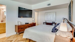 A modern bedroom with a large bed, a TV, a wooden dresser, a desk, and a lamp. - Stylish MCM Retreat: 27 Days or More (Palm Desert)