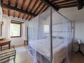 Ein Zimmer mit Himmelbett, einem kleinen Tisch, einem Stuhl, einem Fenster und einer Lampe.