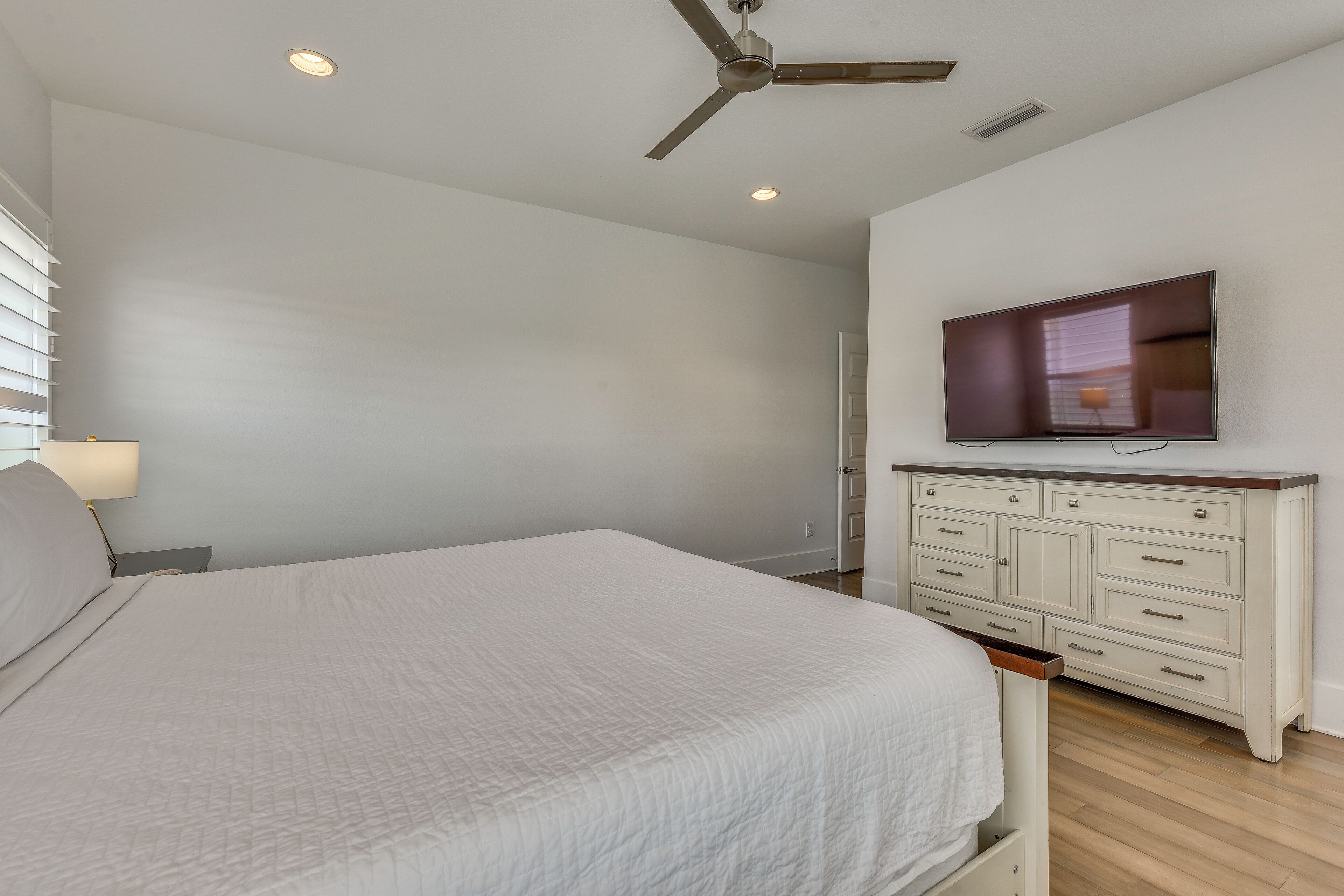 A bedroom with a bed, a TV mounted on a dresser, a ceiling fan, and recessed lighting.