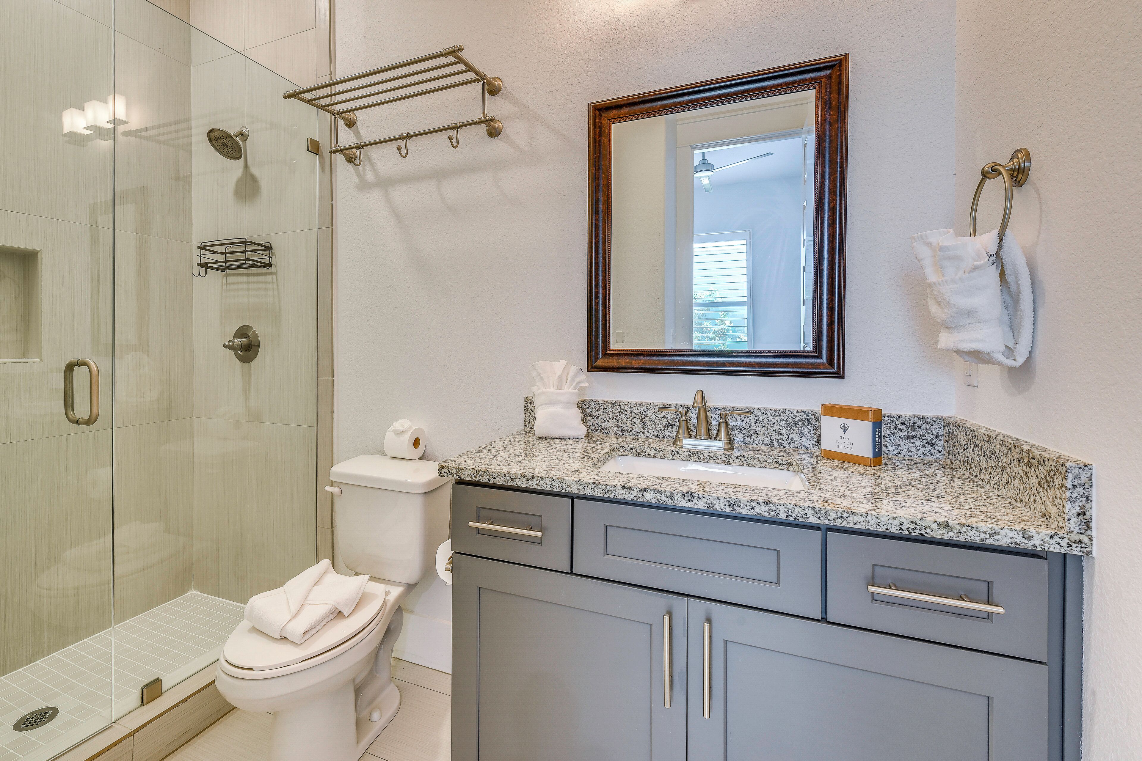 A bathroom with a glass shower enclosure, a toilet, a sink with a granite countertop, and a mirror with a wooden frame.