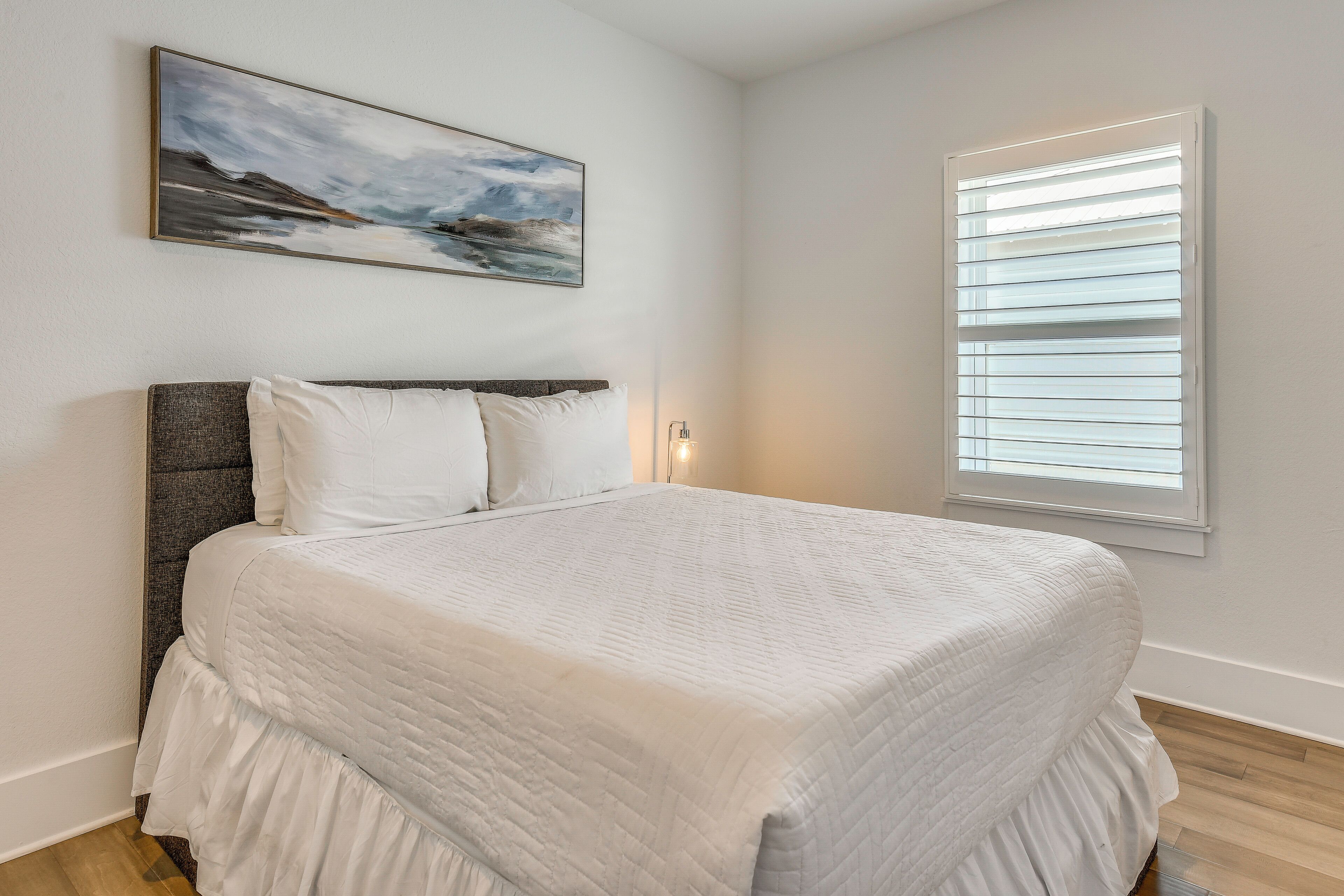 A bedroom with a large bed, a headboard, a painting on the wall, and a window with blinds.